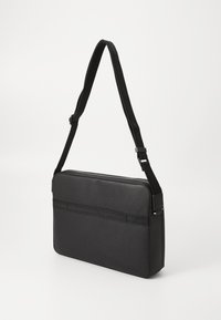 Sac à bandoulière noir avec une surface texturée, de forme rectangulaire et une sangle adjustable. Comprend une fermeture éclair et une poche avant.