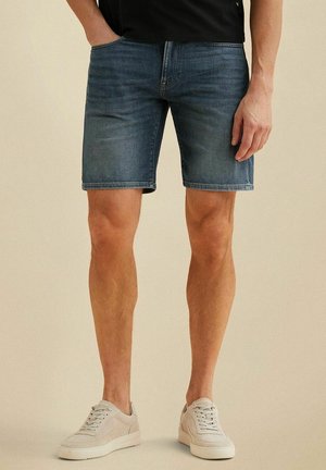 Mann mit blauen Jeansshorts mittlerer Länge und beigen Sneakers, steht mit einer Hand in der Tasche vor einem einfarbigen Hintergrund.
