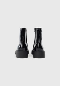 Crne kožne gležnjače s sjajnom površinom, zaobljenim nosom, stražnjim patentnim zatvaranjem i debelim crnim gumiranim potplatom. Glatka tekstura, minimalistički dizajn.