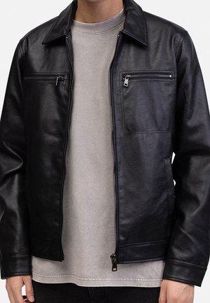 Mann trägt eine schwarze Lederjacke mit Frontreißverschluss und zwei horizontalen, mit Reißverschluss verschließbaren Brusttaschen über einem beigen Hemd.