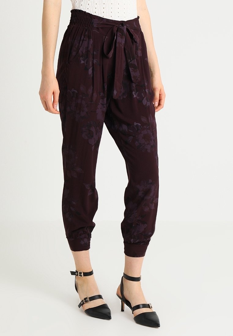 Femme portant un pantalon tapered floral violet foncé avec une taille à nouer et des escarpins noirs à bride de cheville, se tenant devant un fond uni.