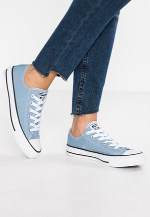 Personne portant des baskets basses en toile bleu clair avec lacets et semelles blanches, associées à un jean bleu foncé à ourlet effiloché.