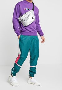 Hombre con sudadera púrpura, pantalones deportivos color teal con rayas rosa y blancas, zapatillas blancas y una mochila cruzada gris.