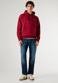 Burgundská mikina s kapucí, přední kapsou a táhly, spárovaná s modrými džíny a béžovými teniskami s černými akcenty.