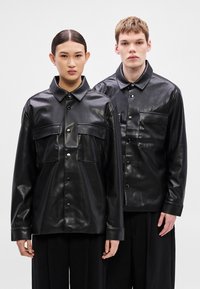 Melnas ādas krekli ar spīdīgu virsmu, kuros ir divas krūšu kabatas un pogu aizdare. Garas piedurknes un klasiskas apkakles dizains.