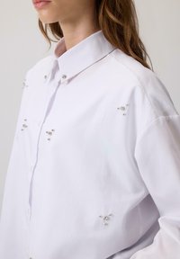 Sieviete, kas valkā baltu krekliņu ar mazām sudraba pērlītēm un rhinestone rotājumiem uz apkakles un priekšpuses.
