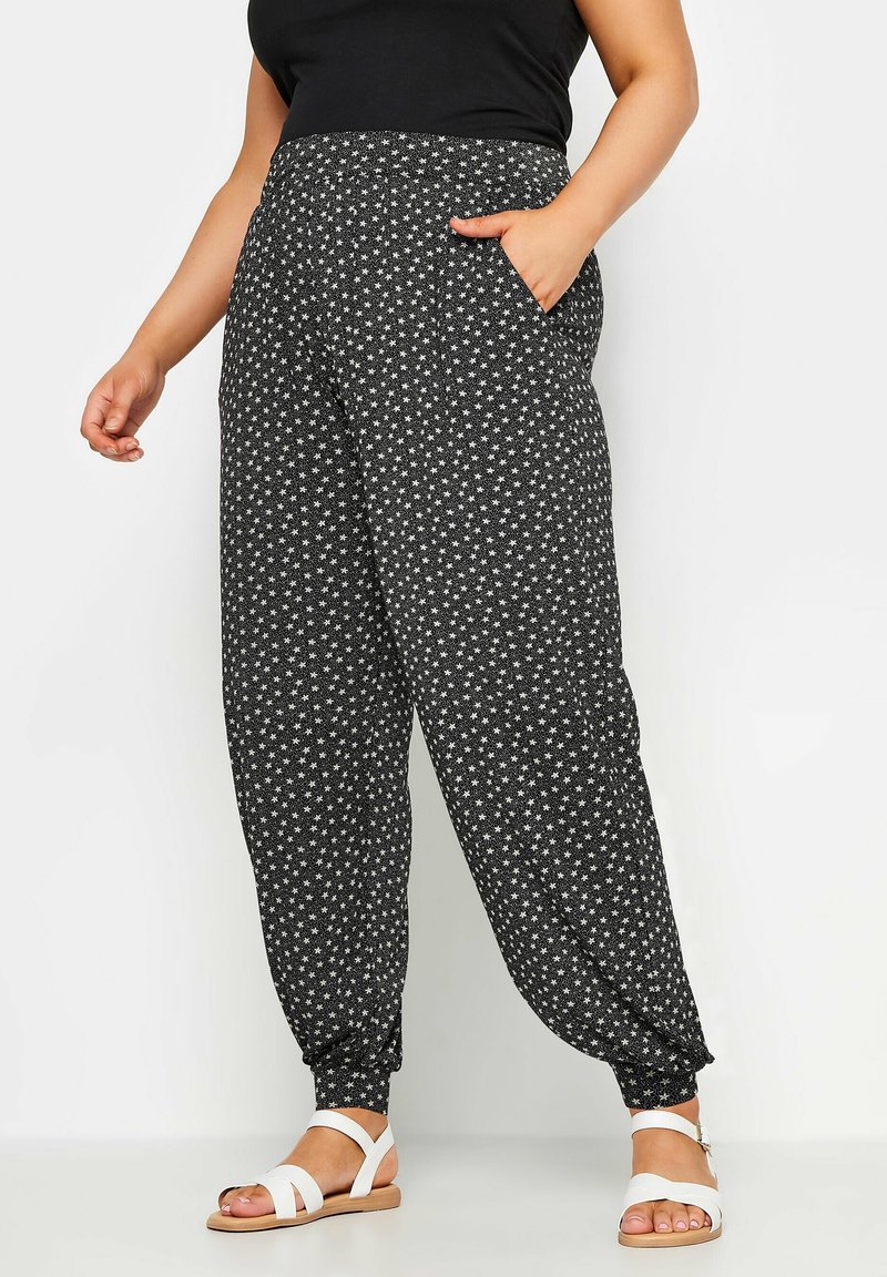 Femme portant un pantalon de jogging noir à motif d'étoiles blanches, associé à des sandales blanches à brides, debout devant un fond clair.