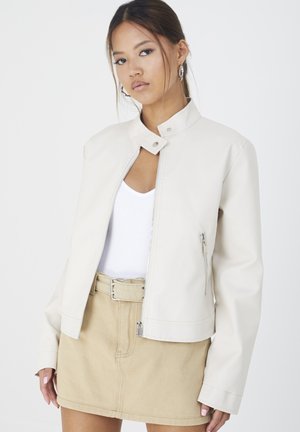 Crème-kleurige faux leren jack met een hoge kraag, ritssluiting aan de voorkant en zijzakken. Gekleed over een witte top en een beige rok.