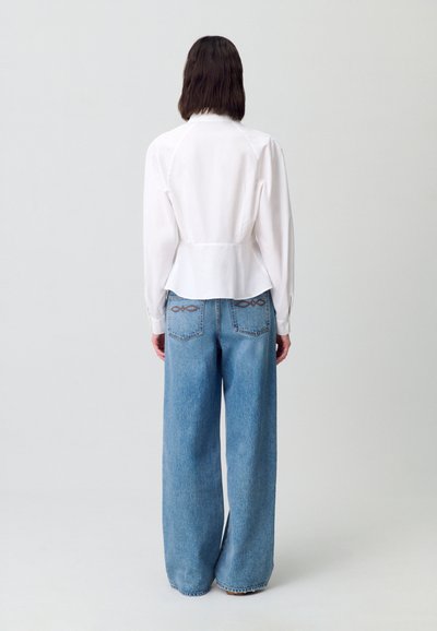 Una persona con capelli scuri fino al mento sta in piedi di spalle, indossando una blusa bianca e jeans a zampa di elefante blu chiaro su uno sfondo semplice.