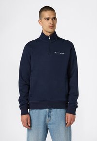 Un uomo che indossa una felpa zip con cappuccio Champion blu navy e jeans blu chiaro, che guarda a sinistra su uno sfondo bianco semplice.