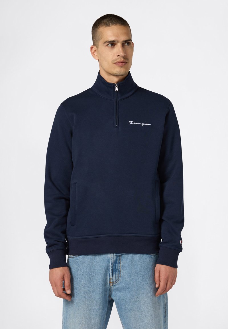 Un uomo che indossa una felpa zip con cappuccio Champion blu navy e jeans blu chiaro, che guarda a sinistra su uno sfondo bianco semplice.