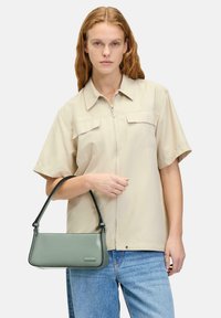 Borsa a mano grigio-verde con un design strutturato e una texture liscia. Presenta una maniglia nera e un sottile dettaglio del marchio.