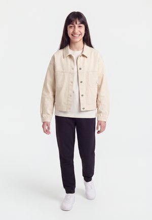 Ung person smiler og går fremad iført en beige jakke, hvid skjorte, sorte bukser og hvide sneakers på en hvid baggrund.
