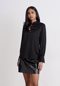 Blusa nera a maniche lunghe con collo a cravatta, abbinata a una mini gonna nera lucida, caratterizzata da trame lisce e silhouette strutturate.