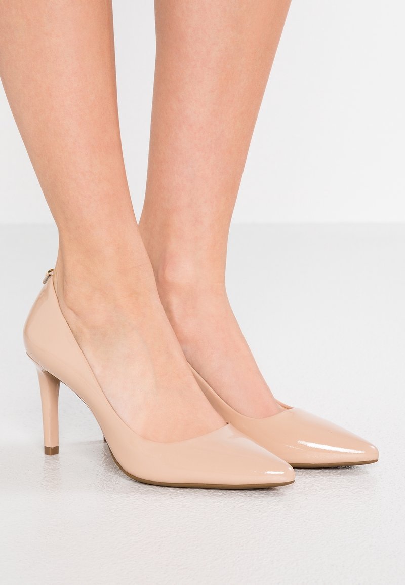Jambes portant des escarpins à bout pointu en cuir verni couleur nude, talons hauts, sur un sol clair avec un fond uni.