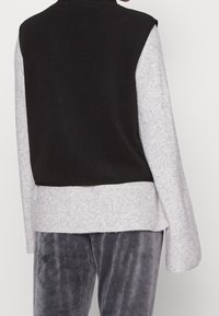 Svart tygväst med hög halsringning, kombinerad med en ljusgrå ribbad långärmad tröja. Mjuk textur och avslappnad passform är synliga.