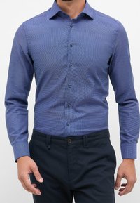 Niebieska koszula w kratę z długim rękawem, zakończona szpiczastym kołnierzykiem i zapinana na guziki z przodu. Noszona z ciemnoniebieskimi spodniami o fakturowanej tkaninie.