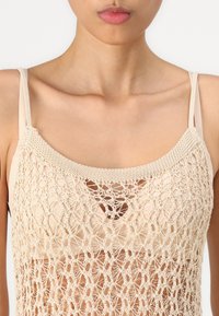 Crochet top in beige, featuring a knitted design with an open weave, adjustable straps, and a rounded neckline.
