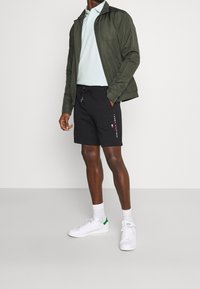 Pantalones cortos negros, cortos, con cordón, chaqueta verde, polo azul claro y zapatillas blancas con acentos verdes. Logo visible en los pantalones cortos.