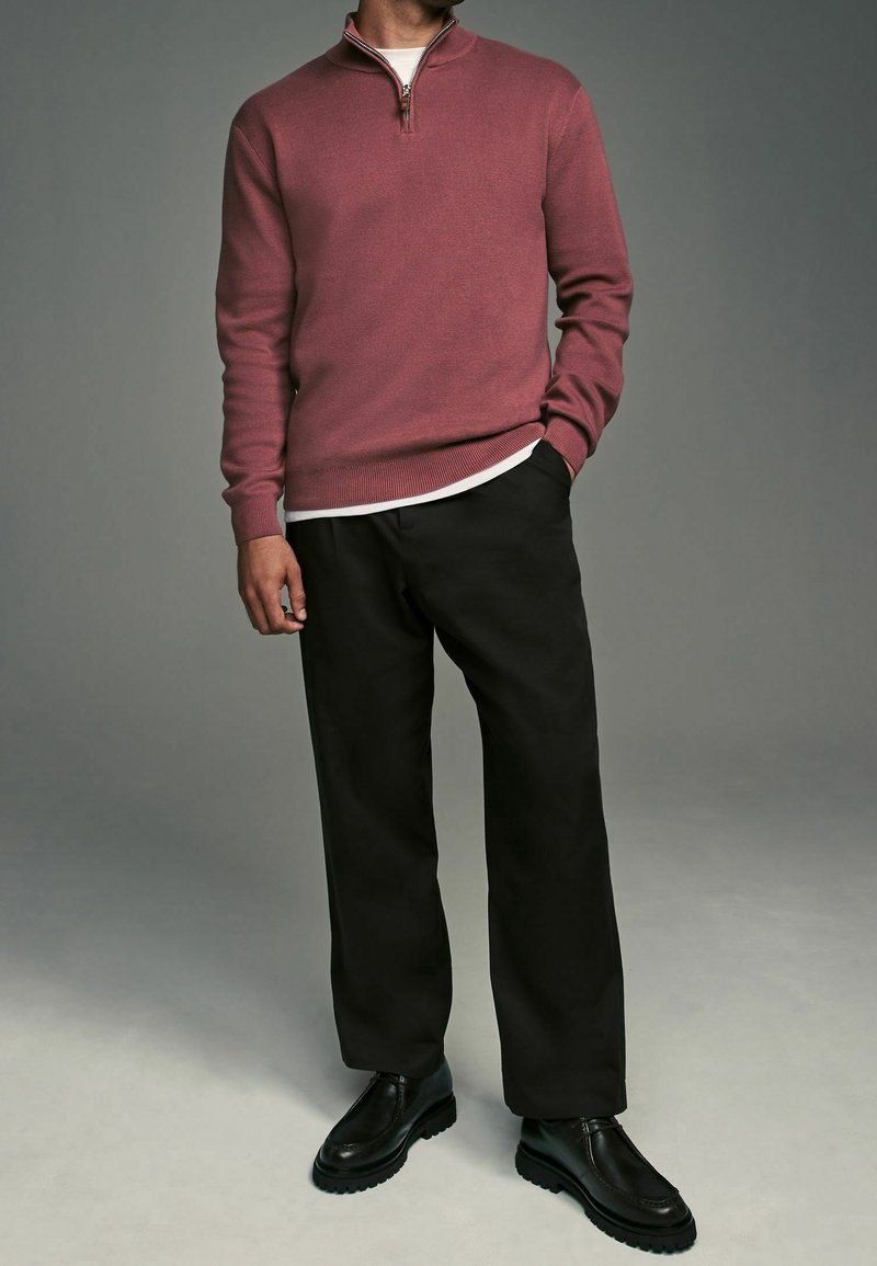 Homme portant un pull rose à fermeture éclair quart, un t-shirt blanc, un pantalon noir et des chaussures en cuir noires, debout avec une main dans la poche.