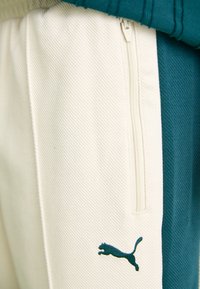 Zelene i kremaste trenirke s dijagonalnom teksturom, s funkcionalnim džepom na zatvarač i tamnozelenim Puma logom na bedru.
