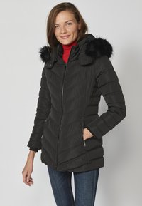 Jachetă puffer neagră cu design quiltat în formă de V, glugă căptușită cu blană, două fermoare laterale și manșete cu dungi. Purtată peste un top roșu.