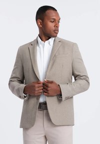 Blazer beige clair en tissu texturé, avec un bouton unique et des revers crantés. Stylé avec une chemise blanche et un pantalon clair.