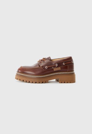 Smeđi kožni loafers s debelim čepastim potplatom, vezice provučene kroz metalne oči, i šivani detalji na bijeloj pozadini.