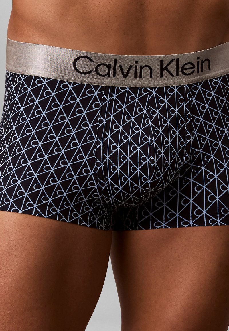 Boxer shorts negros con un patrón geométrico en azul claro, con una cinturilla plateada de contraste que muestra el nombre de la marca.