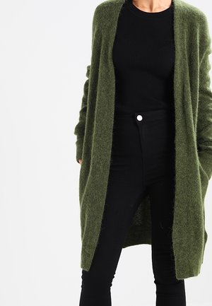 Personne portant un long cardigan duveteux vert olive sur un haut noir côtelé et un pantalon noir taille haute avec un bouton blanc.