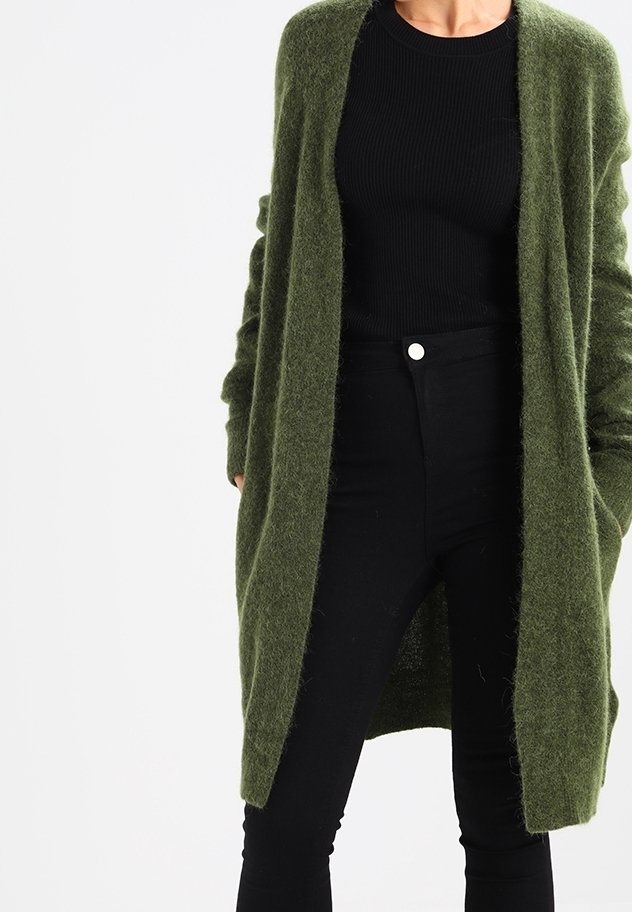 Personne portant un long cardigan duveteux vert olive sur un haut noir côtelé et un pantalon noir taille haute avec un bouton blanc.