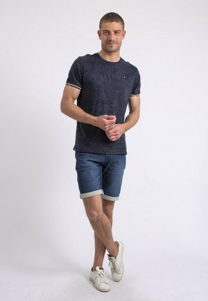 T-shirt marine avec un motif de feuilles texturé, des poignets contrastants, associé à un short en denim avec des ourlets retroussés et des baskets blanches.