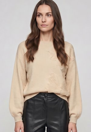 Frau mit langen braunen Haaren, die einen beigen Pullover mit Blumenstickerei und schwarze Lederhose trägt, steht vor einem schlichten Hintergrund.