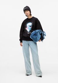 Melnā džemperī ar krāsainu grafiku, gaiši zila plats kāju bikses un zila auduma soma. Modelis valkā melnu cepuri ar logotipu.