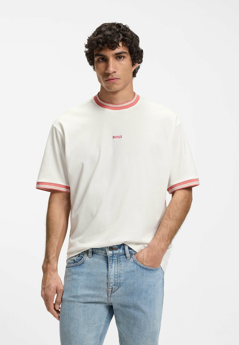 Jeune homme aux cheveux bouclés portant un t-shirt blanc à bordure rouge avec le logo "BOSS", associé à un jean bleu clair, debout devant un fond uni.