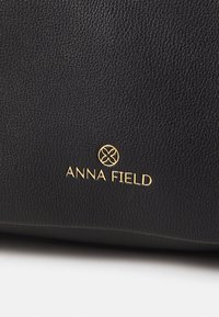 Borsa a tracolla in pelle nera con superficie testurizzata e logo dorato "ANNA FIELD" con un accento circolare, che mette in evidenza il nome del marchio.