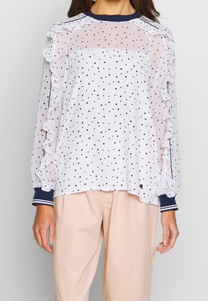 Blouse blanche transparente à manches longues avec des pois noirs et des volants le long des manches, associée à un pantalon beige clair.