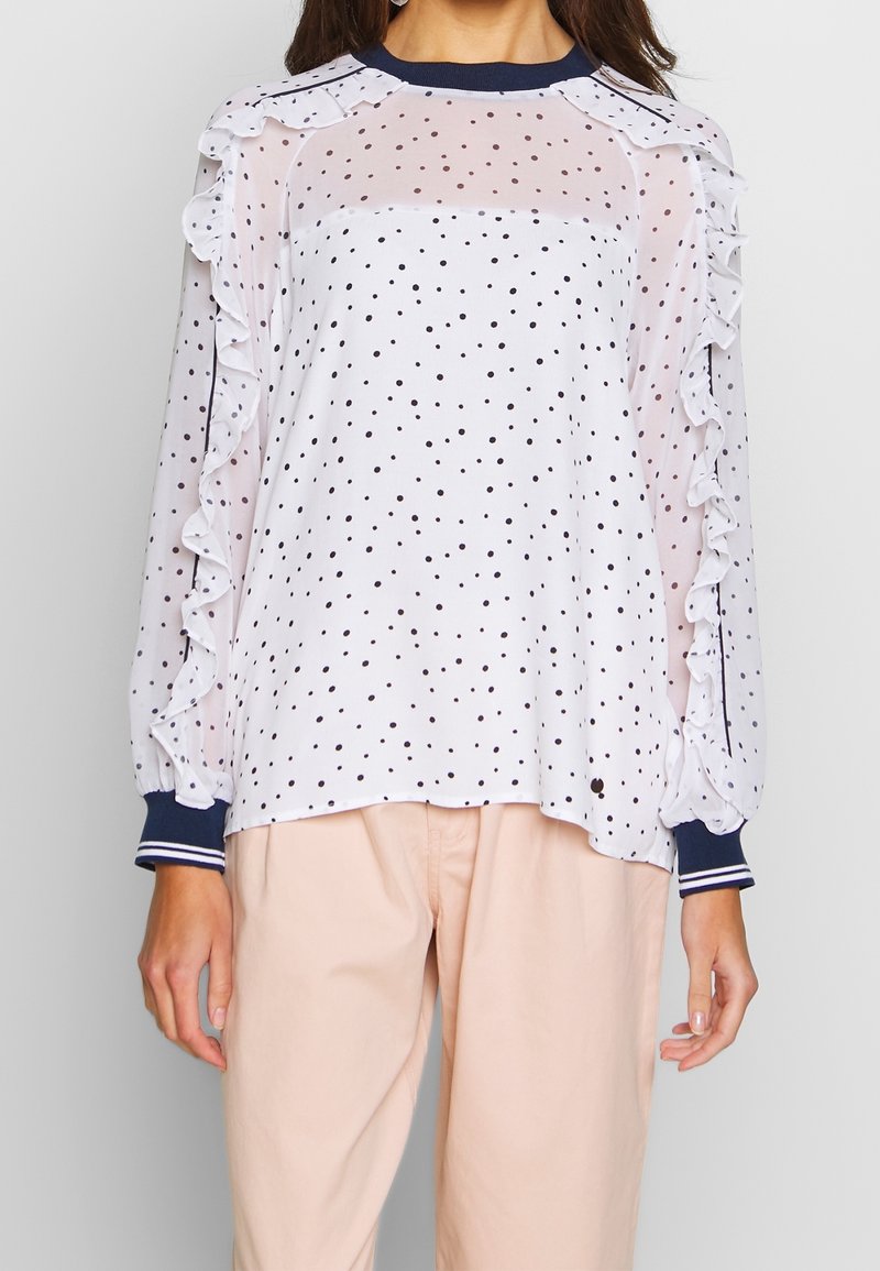 Blouse blanche transparente à manches longues avec des pois noirs et des volants le long des manches, associée à un pantalon beige clair.