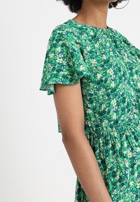 Grön klänning med blommönster, korta volangärmar, rund halsringning och rynkad midja, tillverkad av lättviktig tyg.