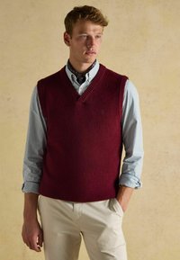 Burgundowa wełniana kamizelka bez rękawów z dekoltem w serek, noszona na jasnoniebieskiej i białej w paski koszuli z guzikami oraz kremowymi spodniami.