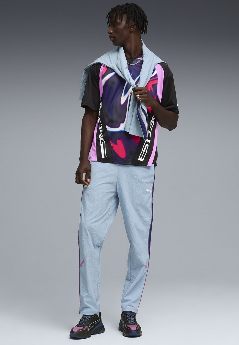 Jeune homme portant un t-shirt graphique noir et rose, un pantalon de survêtement bleu clair, des baskets noires et roses, avec un sweat-shirt bleu clair drapé sur les épaules.