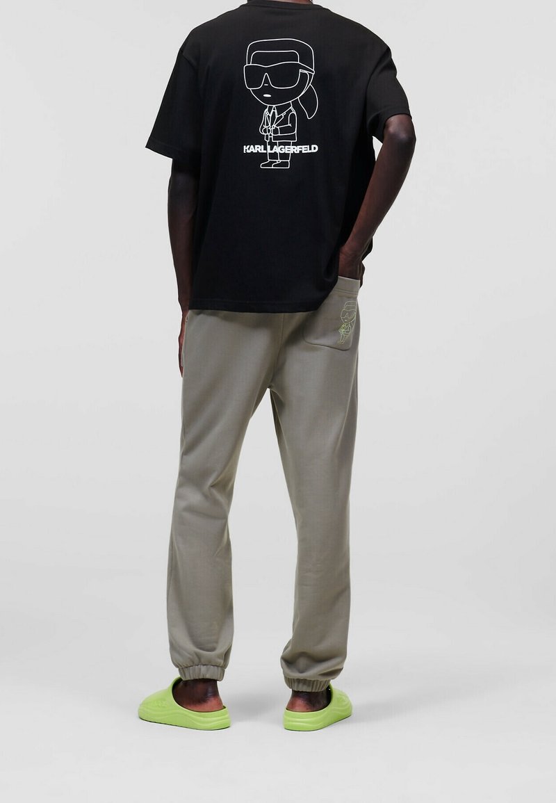 Černé tričko s karikaturou a bílým textem na zádech, doplněné šedými tepláky a jasně zelenými nazouvacími botami.