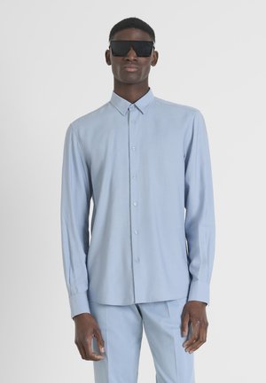 Lichtblauw overhemd met een kraag, lange mouwen en een gladde textuur, gedragen met bijpassende lichtblauwe broek. Zwarte zonnebril gedragen.