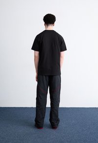 Zwarte katoenen T-shirt en losse zwarte broek met rode stikdetails, gezien vanaf de achterkant tegen een egale wand en een vloer met tapijt.