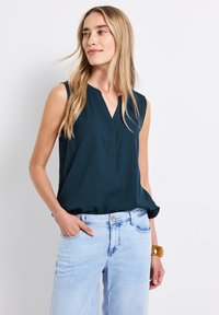 Blouse sans manches en vert foncé à décolleté en V, en tissu doux, avec des détails de couture subtils, associée à un jean en denim bleu clair. Accessoire bracelet en bois.