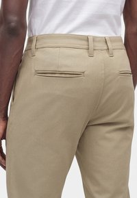 Homme portant un pantalon beige avec des poches passepoilées à l’arrière et des passants de ceinture, associé à une chemise blanche, présenté de la taille jusqu’au milieu de la cuisse.