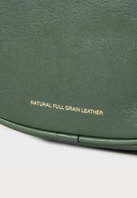 Pelle di vera pelle pieno fiore verde naturale con una superficie testurizzata. Presenta testo dorato embossato: "PELLA DI VERA PELLE PIENO FIORE." Forma rotonda.