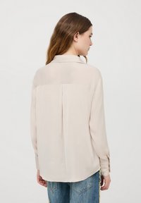 Blusa beige chiaro con maniche lunghe e orlo curvo. Presenta una piega sul retro e un colletto classico. Realizzata in tessuto morbido e leggero.