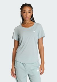 Žena s pletenými vlasmi, oblečená v svetlomodrom tričku s krátkym rukávom Adidas a zodpovedajúcich nohaviciach na jednoliatom pozadí.