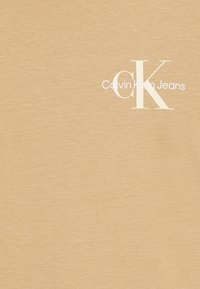 Világos bézs pamut póló, amelyen előlapján egy kis fehér "Calvin Klein Jeans" logó és "CK" dizájn található, sima textúrával.